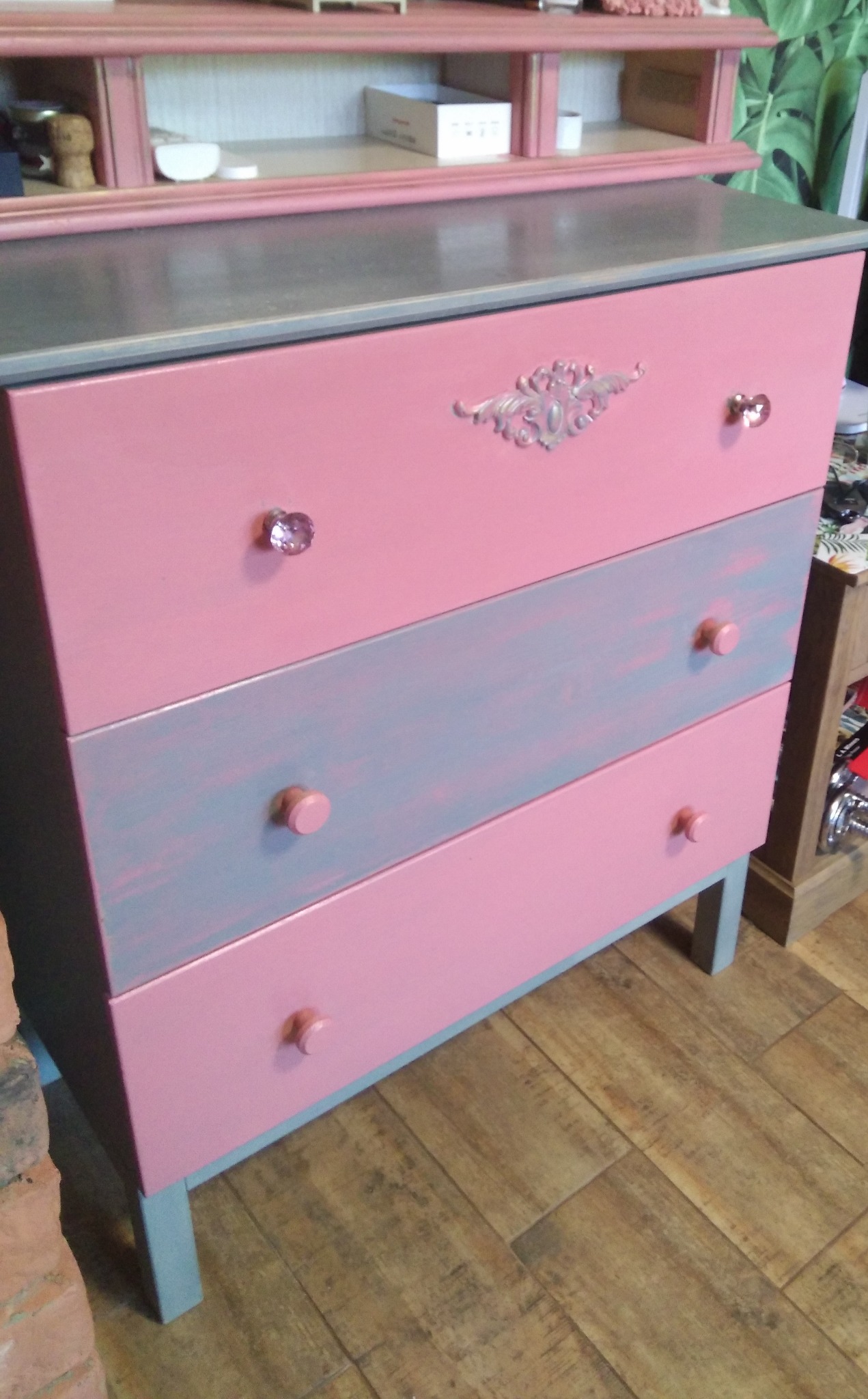 Komoda z trzema szufladami w odcieniach różu i szaro-niebieskiego, z dekoracyjnym ornamentem i kryształowymi gałkami, stojąca na drewnianej podłodze.