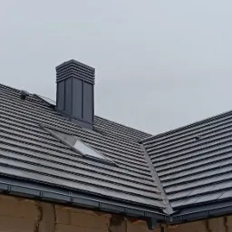 Nowoczesny dach z dachówki płaskiej Piano w kolorze antracytowym, z oknami połaciowymi Velux i systemem rynnowym Galeco, na budynku w trakcie budowy z widoczną konstrukcją drewnianą i murowanymi...