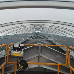 Wnętrze hali w trakcie budowy, widok z pomostu roboczego na stalową konstrukcję dachu z białym, półprzezroczystym pokryciem i panelem sterowania.