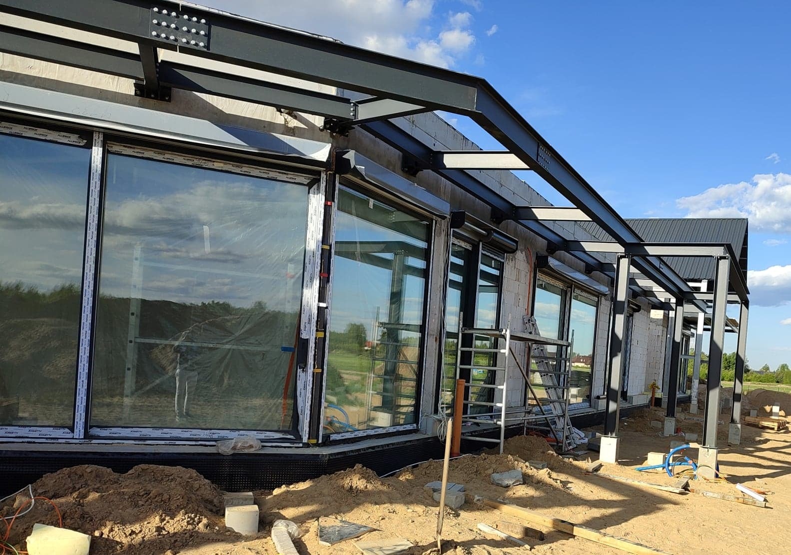 Konstrukcja stalowa zadaszenia tarasu przy budynku w trakcie budowy, widoczne duże okna z folią ochronną i rusztowanie.