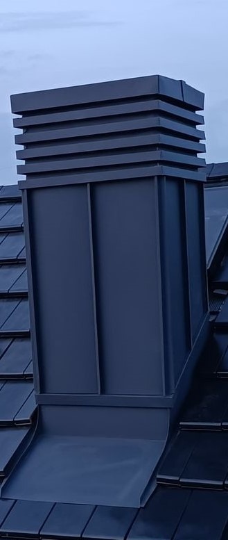 Nowoczesny, ciemnoszary komin z obróbką blacharską, wentylowany, na dachu pokrytym ciemną dachówką.