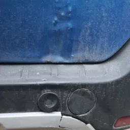 Wgniecenie i odprysk lakieru na brudnym, niebieskim błotniku samochodu, widoczny uszkodzony, srebrny fragment zderzaka z czujnikami parkowania.