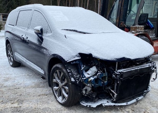 Uszkodzony, granatowy minivan pokryty śniegiem, z widocznymi uszkodzeniami przodu i odsłoniętą chłodnicą, stojący na ośnieżonym placu w zimowy dzień.
