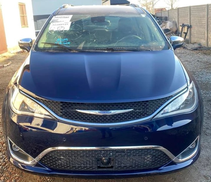 Niebieski samochód Chrysler Pacifica z uszkodzoną przednią szybą i notatkami na niej, stojący na zewnątrz przed betonowym ogrodzeniem.