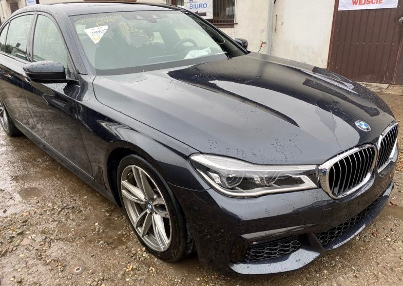 Ciemny, mokry samochód BMW serii 7, zaparkowany przed budynkiem z napisem 'BIURO' i 'WEJŚCIE', z naklejką na przedniej szybie.