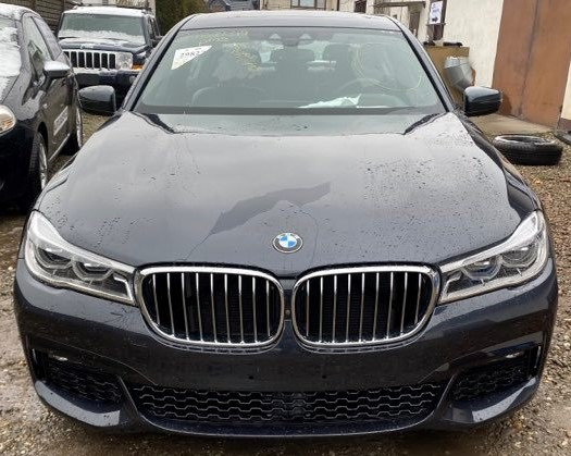 Przód ciemnoszarego samochodu BMW serii 7 z widocznymi kroplami wody na karoserii, stojącego na żwirowym podłożu, z innymi pojazdami w tle.