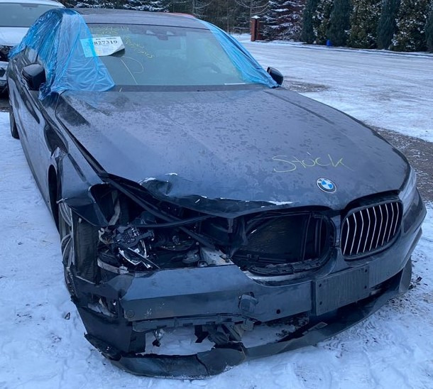 Uszkodzony przód ciemnego samochodu BMW, widoczne zniszczenia karoserii i brakujące elementy, na masce foliowa osłona, na śniegu.