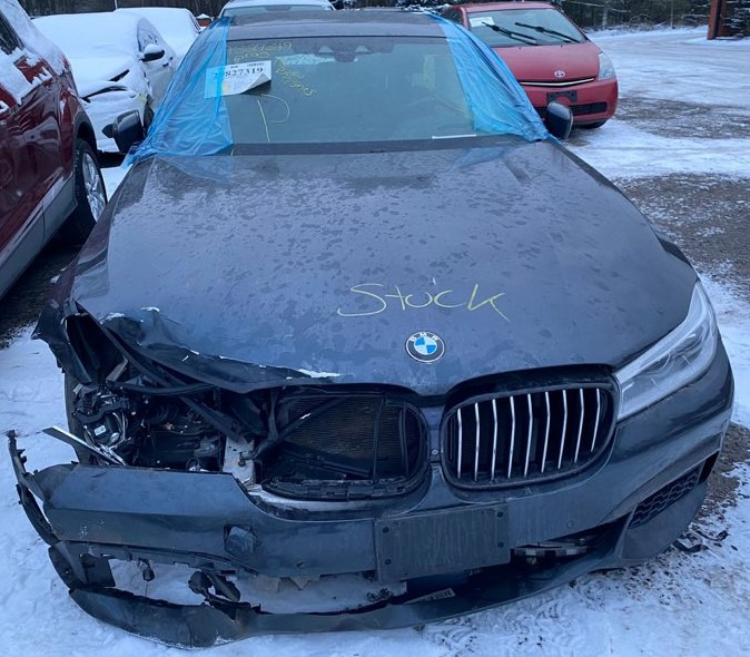 Uszkodzony przód ciemnoszarego samochodu BMW na parkingu, widoczne wgniecenia i pęknięcia karoserii, zarysowane napisem 'Stock' na masce, z niebieską folią na przedniej szybie.