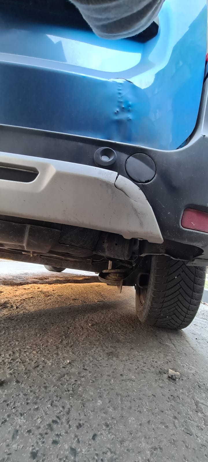 Uszkodzony, wgnieciony tylny błotnik niebieskiego samochodu, widoczne pęknięcie na szarym zderzaku, ujęcie z dołu.