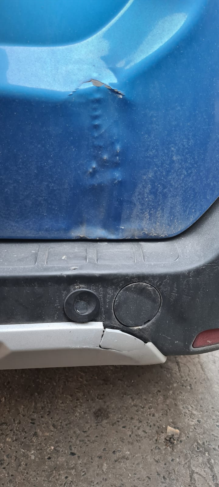 Wgniecenie i odprysk lakieru na brudnym, niebieskim błotniku samochodu, widoczny uszkodzony, srebrny fragment zderzaka z czujnikami parkowania.