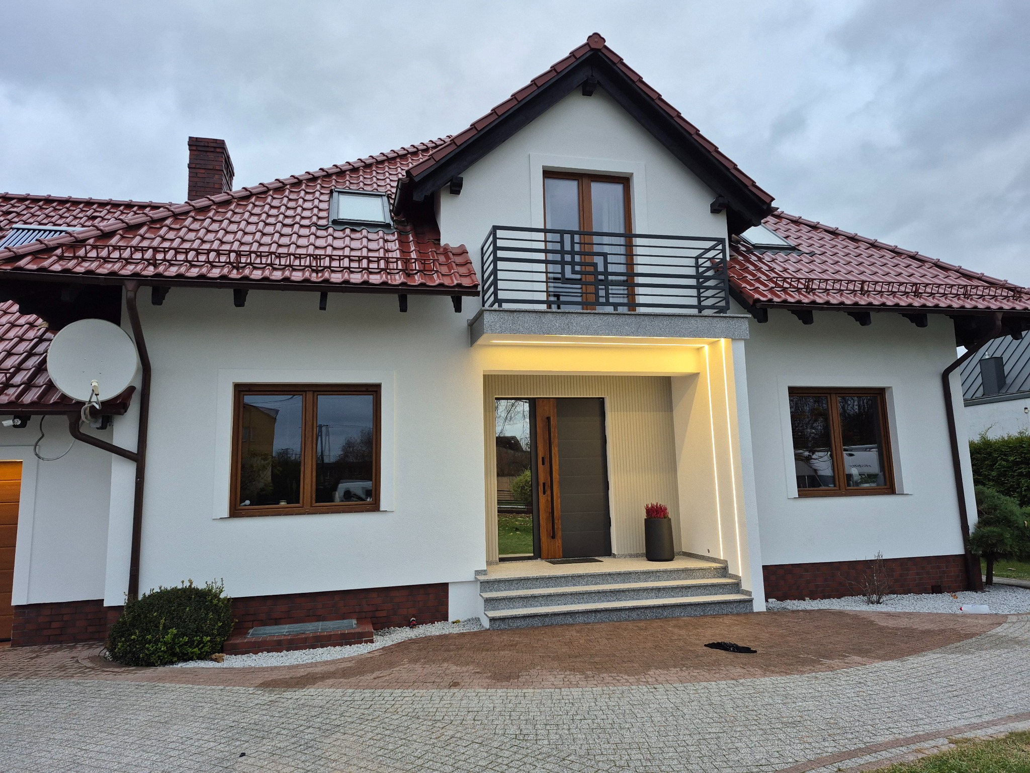 Nowoczesny dom jednorodzinny z białą elewacją, czerwonym dachem, balkonem z czarną balustradą i podświetlanym wejściem, widok z poziomu brukowanego podjazdu.