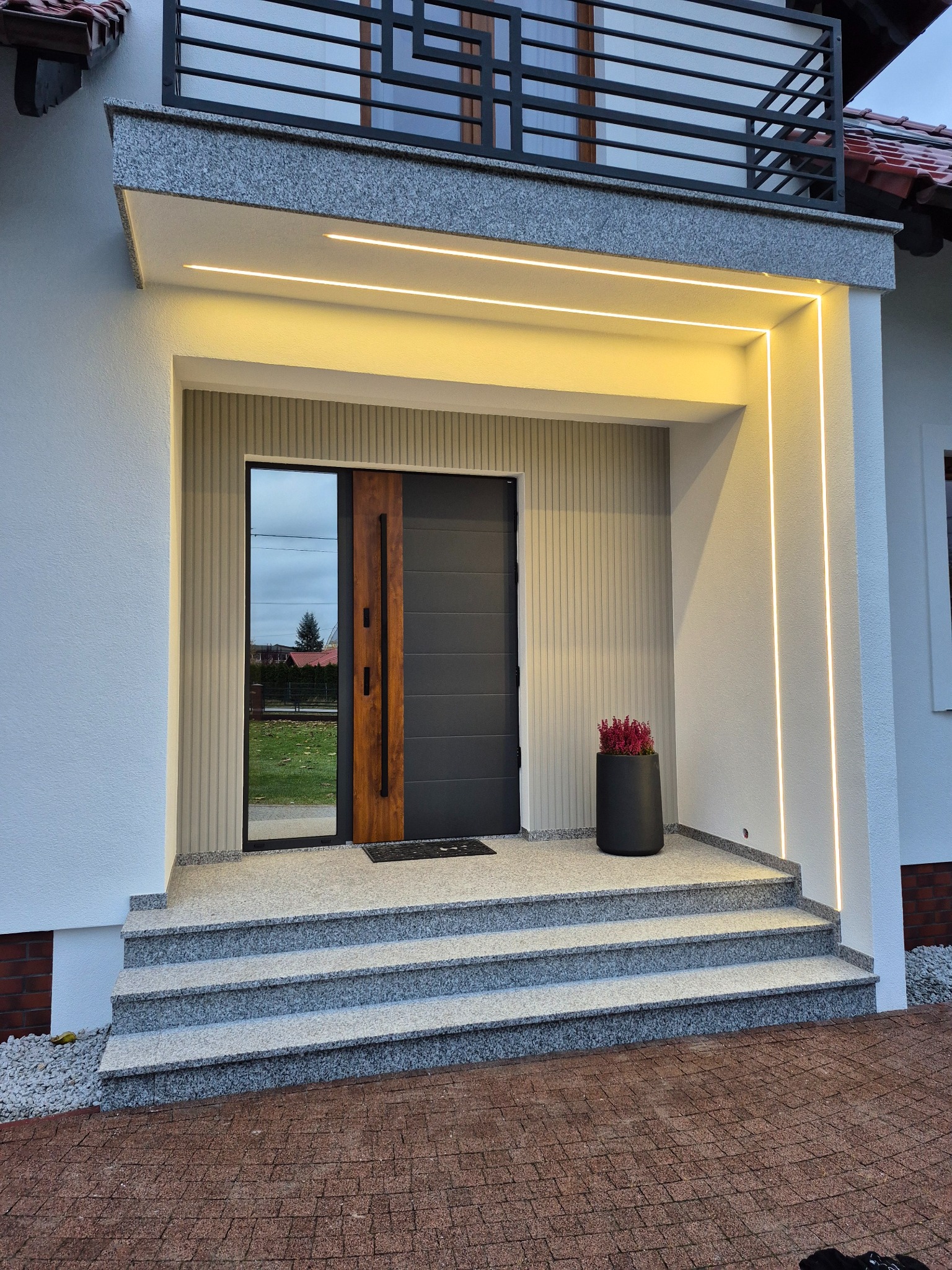 Nowoczesne wejście do domu z szarymi schodami granitowymi, ciemnymi drzwiami z drewnianym akcentem i oświetleniem LED wzdłuż elewacji, uzupełnione czarną donicą z czerwoną rośliną.