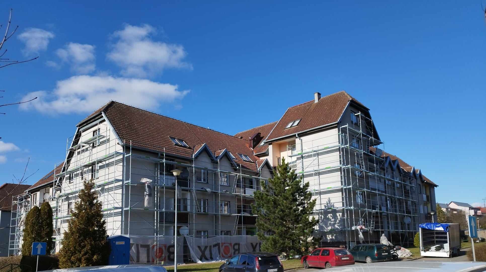 Wielopiętrowy budynek mieszkalny w trakcie prac elewacyjnych, otoczony rusztowaniami, widoczne osoby pracujące na rusztowaniach, błękitne niebo z chmurami w tle.