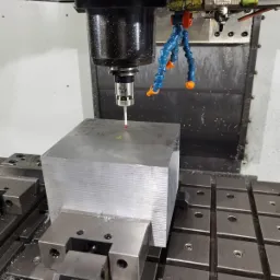 Blok metalu zamocowany w uchwycie maszyny CNC, pomiar punktu sondą pomiarową, widoczne dysze chłodziwa.