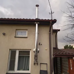 Zewnętrzny, stalowy przewód spalinowy zamontowany na elewacji domu z numerem 4A, widoczny drabina oparta o ścianę.