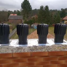 Cztery czarne, nowe nasady kominowe zamontowane na ceglanym kominie, zabezpieczone srebrną folią uszczelniającą, w tle zabudowa jednorodzinna i drzewa.