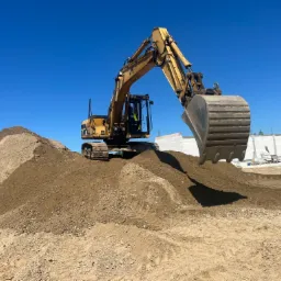 Żółta koparka na szczycie usypanej ziemi, z widocznym operatorem w kabinie, na tle błękitnego nieba i częściowo widocznego budynku w budowie.