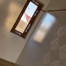 Nowoczesna łazienka z białymi, błyszczącymi płytkami, oknem dachowym z brązową ramą i złotą deszczownicą, w tle widoczny fragment drewnianej okładziny ściany.