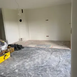 Przygotowanie do malowania ścian w Szprotawie: zabezpieczona folią podłoga, puszki z farbą i narzędzia malarskie w pustym pomieszczeniu z instalacją elektryczną.