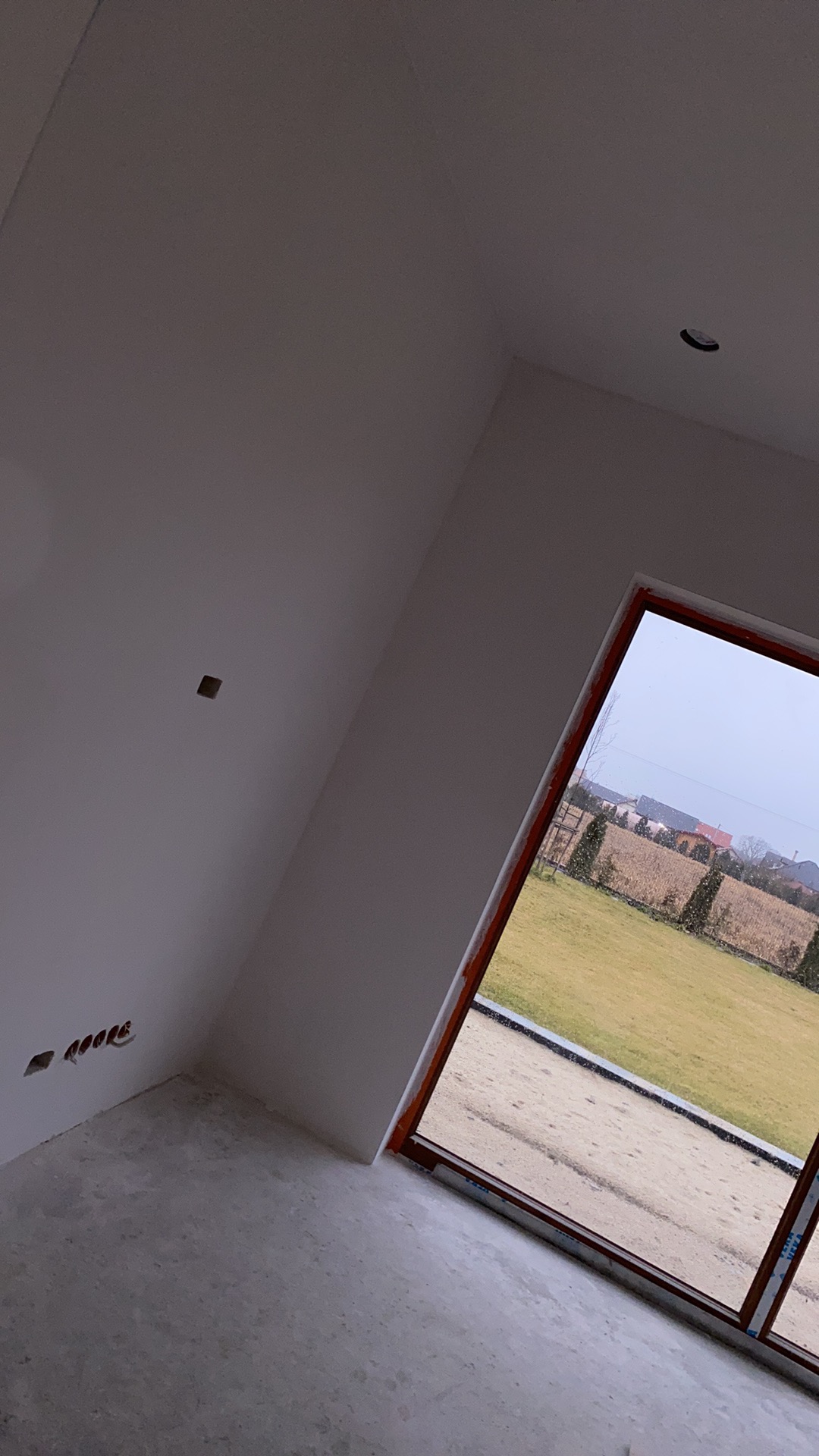 Surowe wnętrze pokoju z oknem na zewnątrz, ściany przygotowane pod wykończenie, widoczne otwory na gniazdka elektryczne i surowy beton na podłodze, za oknem trawnik i domy.