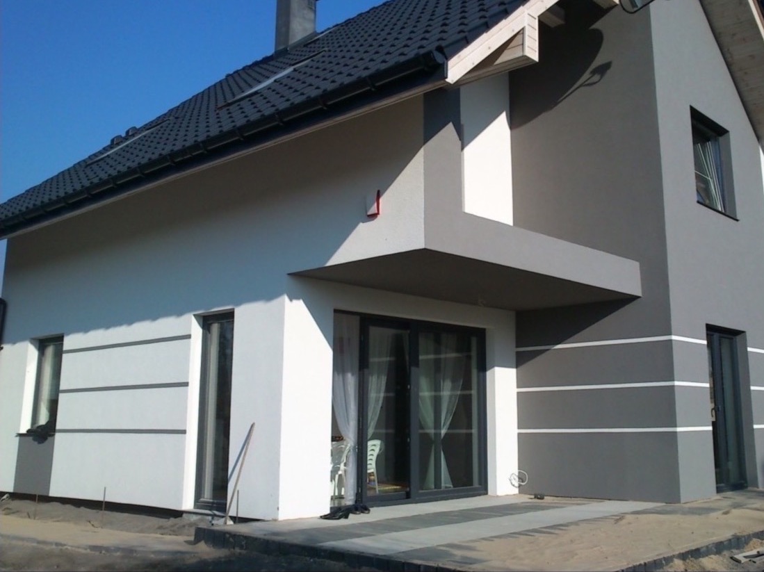 Dwukolorowa elewacja domu jednorodzinnego z grafitową dachówką i szarymi ramami okiennymi, widok z zewnątrz na fragment budynku z widocznymi detalami wykończenia.