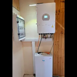 Biała bateria magazynu energii, inwerter solarny Deye i skrzynka bezpiecznikowa zamontowane na ścianie w pomieszczeniu z drewnianą boazerią.