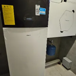 Kompaktowa instalacja grzewcza Daikin z widocznymi przewodami wentylacyjnymi, niebieskim zbiornikiem wyrównawczym i szarą skrzynką elektryczną na tle szarej płytki ceramicznej.