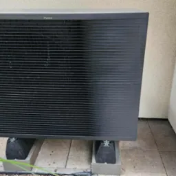 Szara jednostka zewnętrzna klimatyzacji Daikin, ustawiona na betonowych blokach na zewnątrz budynku z jasną elewacją i roślinnością w tle.