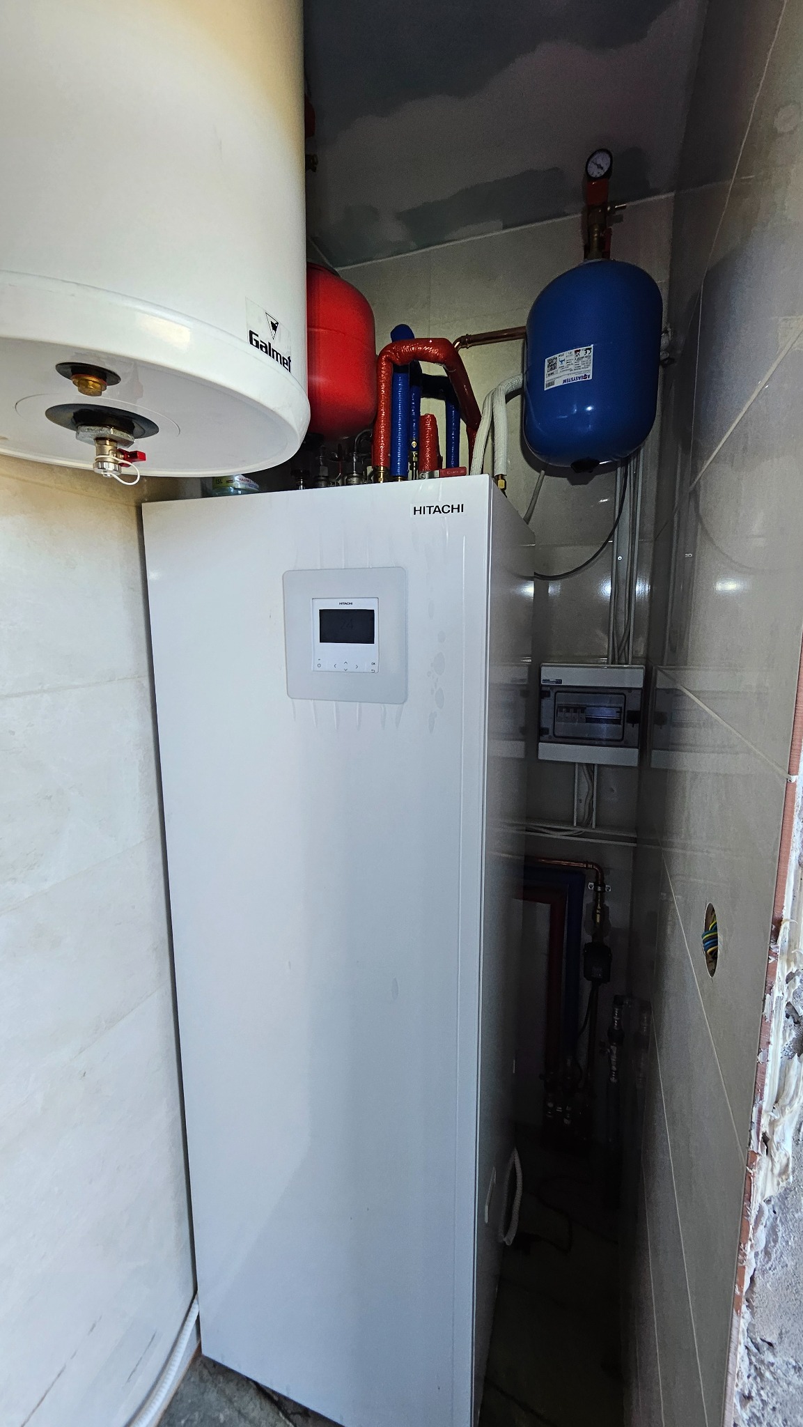 Biała pompa ciepła Hitachi z wyświetlaczem, zbiornik Galmet u góry, czerwony i niebieski zbiornik wyrównawczy, instalacja hydrauliczna z rurami w izolacji, szara skrzynka elektryczna na ścianie...