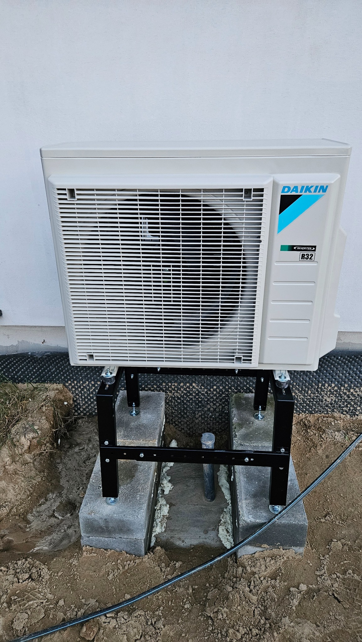 Biała jednostka zewnętrzna pompy ciepła Daikin Inverter R32, zamontowana na czarnej metalowej konstrukcji na betonowych bloczkach, widoczne uszczelnienia pianką montażową i wystająca rura odpływowa.