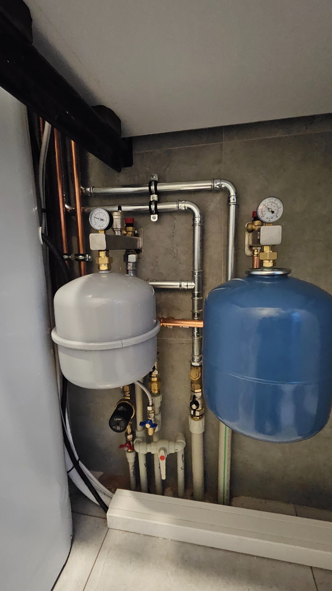 Dwa zbiorniki wyrównawcze, szary i niebieski, z manometrami i rurami miedzianymi oraz z tworzywa sztucznego, zamontowane na szarej ścianie w pomieszczeniu technicznym.