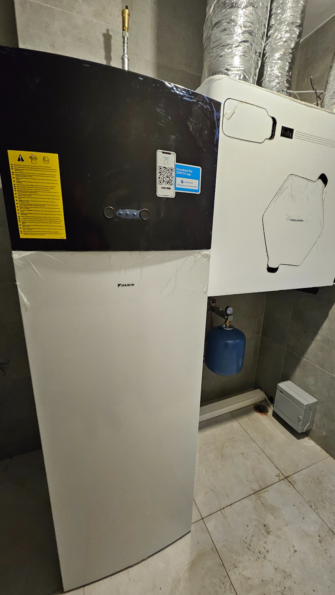 Kompaktowa instalacja grzewcza Daikin z widocznymi przewodami wentylacyjnymi, niebieskim zbiornikiem wyrównawczym i szarą skrzynką elektryczną na tle szarej płytki ceramicznej.