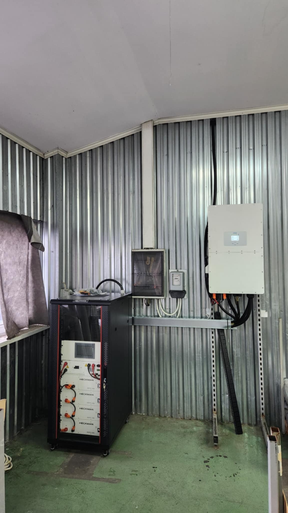 Wnętrze pomieszczenia gospodarczego z zainstalowanym systemem fotowoltaicznym: inwerter na ścianie, obok skrzynka elektryczna i licznik, oraz szafa rack z akumulatorami Heckman.