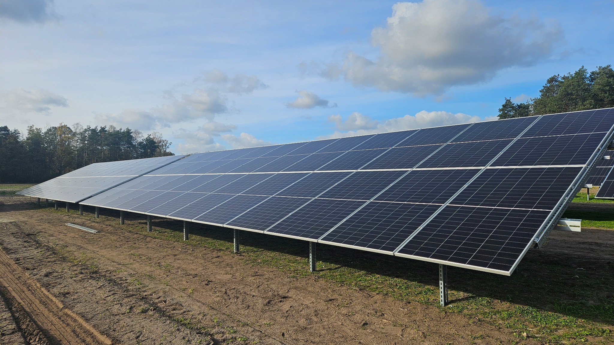 Rząd paneli fotowoltaicznych zamontowanych na metalowych konstrukcjach na polu z widocznymi śladami opon, w tle las i błękitne niebo z chmurami.