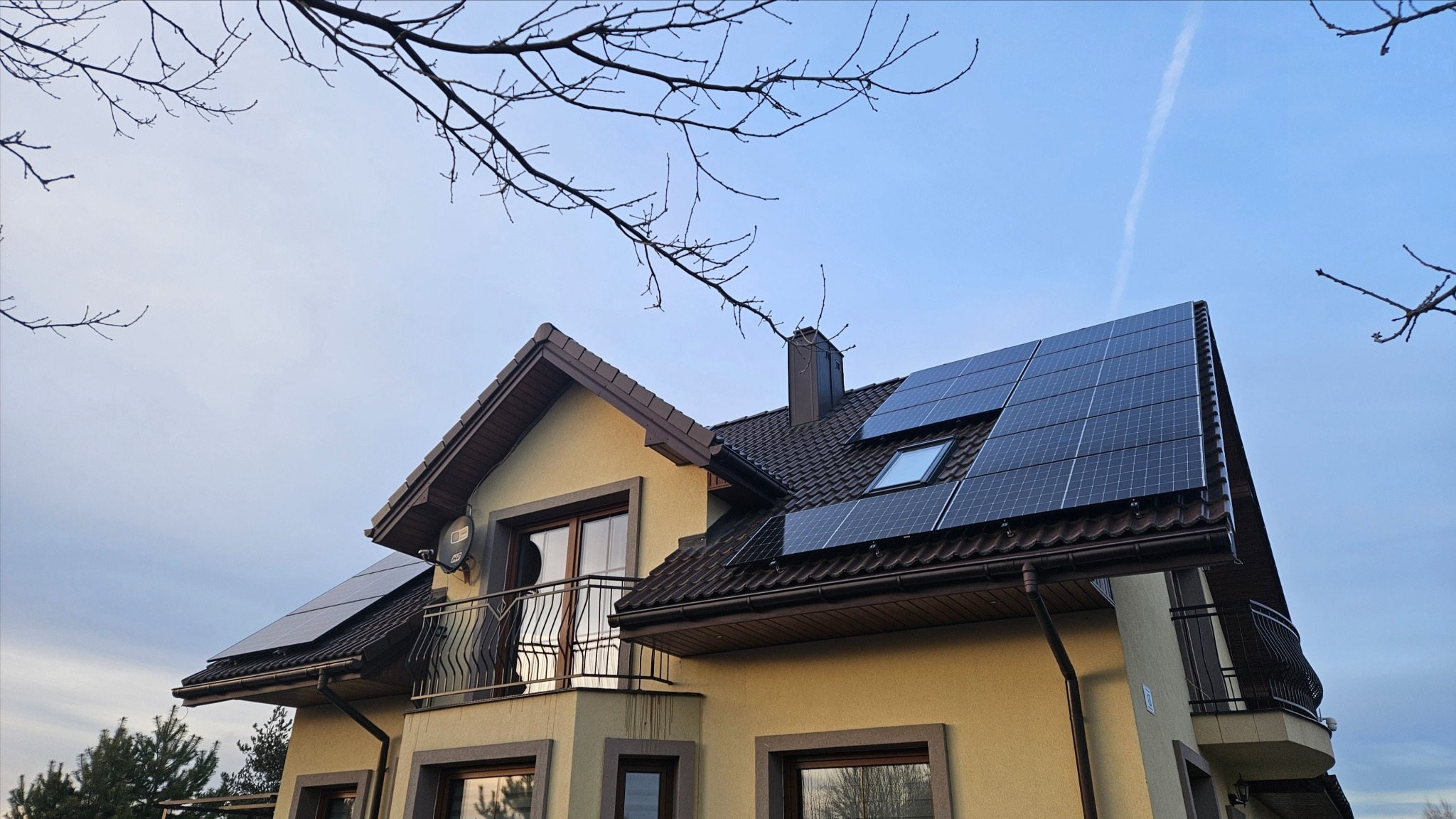 Dwukondygnacyjny dom z beżową elewacją i brązowym dachem, na którym zamontowano panele słoneczne; widoczny komin i balkon z metalową balustradą, na tle błękitnego nieba i gałęzi drzew
