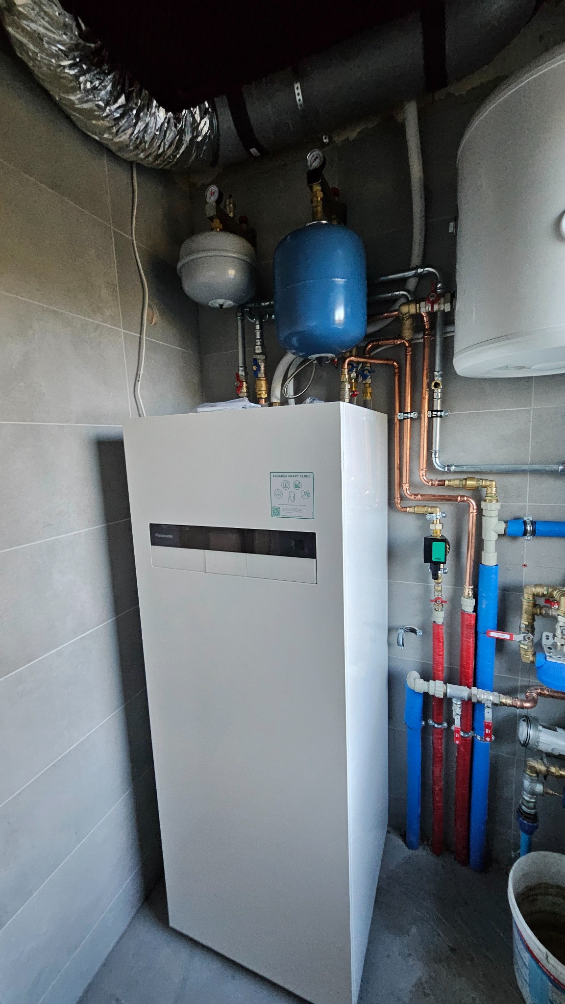 Biała pompa ciepła Panasonic Aquarea Smart Cloud z niebieskim zbiornikiem wyrównawczym i instalacją hydrauliczną z rur miedzianych i PCV w pomieszczeniu z szarymi płytkami.