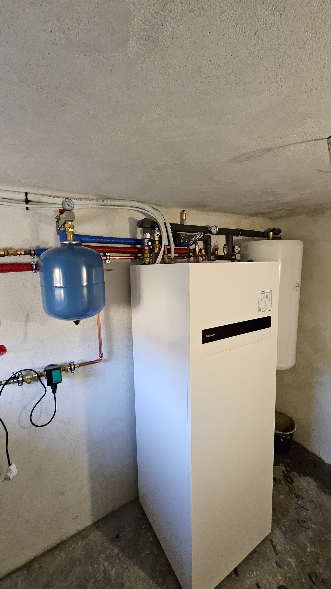 Biała jednostka wewnętrzna z zbiornikiem buforowym, instalacja rurowa w kolorach czerwonym i niebieskim, zamontowana w piwnicy w Łasku.