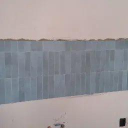 Ściana w trakcie kładzenia niebieskich, pionowych płytek w łazience, widoczne czarne gniazdka elektryczne i ślady po zaprawie na górze ściany.