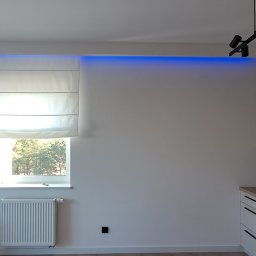 Dizajn Jarosław Lauer - Nowoczesna kuchnia z białymi szafkami, drewnianym blatem, czarną płytą grzewczą, marmurowym backsplash, oknem z roletą rzymską i dekoracyjną ścianą z paneli drewnianych podświetlonych niebieskim...