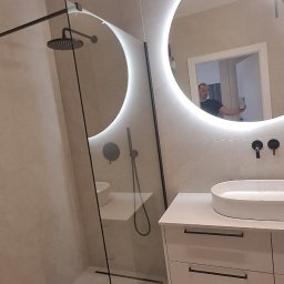 Dizajn Jarosław Lauer - Nowoczesna łazienka z kabiną prysznicową typu walk-in, okrągłym lustrem z podświetleniem LED i białą szafką z umywalką nablatową. Widoczne czarne baterie.