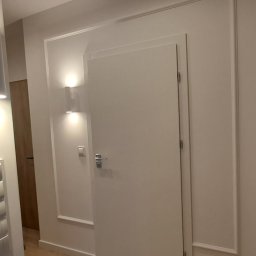 Dizajn Jarosław Lauer - Białe drzwi wewnętrzne z ozdobnymi listwami, oświetlenie punktowe na ścianie, widok na fragment korytarza z drewnianymi drzwiami w tle. Minimalistyczny design.