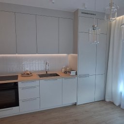 Dizajn Jarosław Lauer - Jasna kuchnia z szarymi szafkami, drewnianym blatem i trzema szklanymi lampami wiszącymi. Minimalistyczny design z białymi zasłonami i drewnianą podłogą.