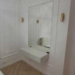 Dizajn Jarosław Lauer - Elegancka toaletka z lustrem w białej ramie, zamontowana na ścianie w pokoju z parkietem ułożonym w jodełkę. Dwa złote kinkiety po bokach lustra.