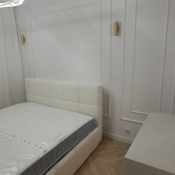 Dizajn Jarosław Lauer - Biała sypialnia z łóżkiem tapicerowanym i panelami ściennymi. Złote kinkiety oświetlają przestrzeń. Podłoga w jodełkę. Nowoczesny design.