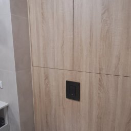Dizajn Jarosław Lauer - Nowoczesna toaleta podwieszana z zabudową meblową w odcieniach drewna. Czarny przycisk spłuczki na tle drewnianej ściany. Minimalistyczny design łazienki.