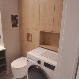 Dizajn Jarosław Lauer - Jasna łazienka z drewnianą zabudową meblową, toaletą podwieszaną i pralką z umywalką na blacie. Minimalistyczny design, neutralna kolorystyka.