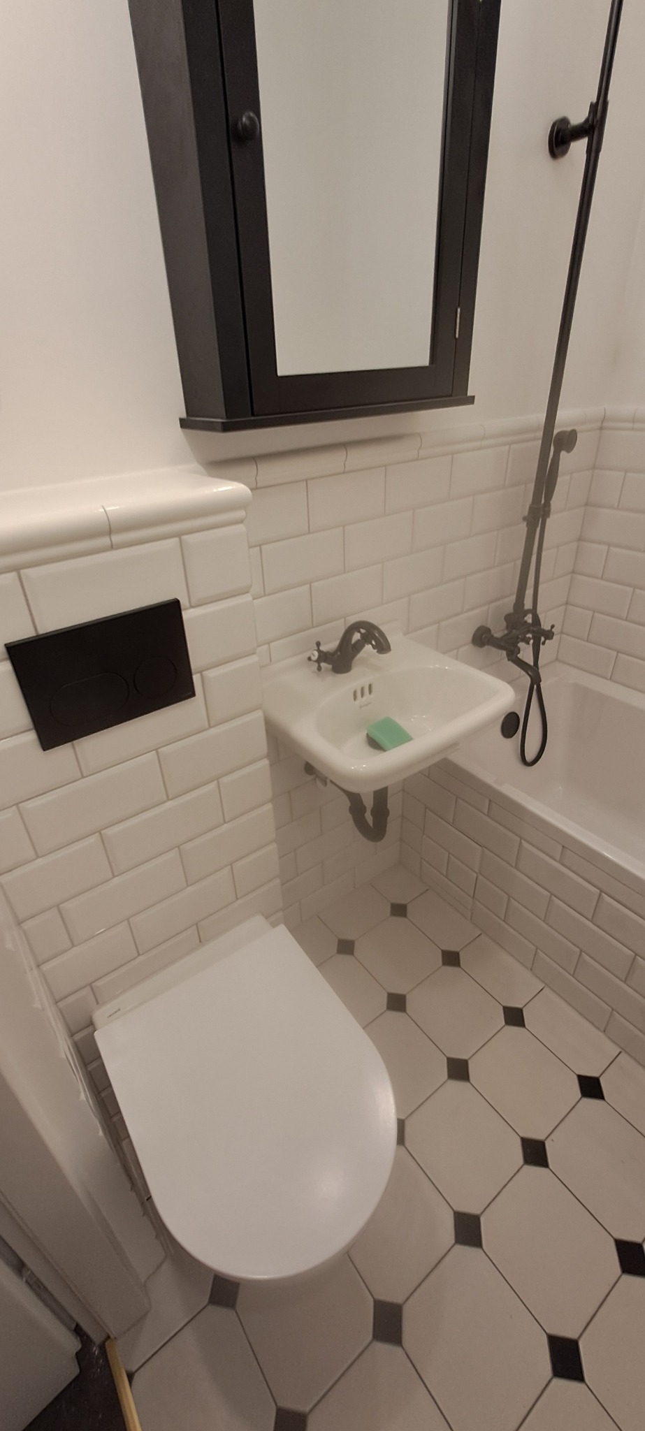 Biała łazienka z czarnymi akcentami: szafka nad umywalką, bateria, przycisk spłuczki i wzór na podłodze. Umywalka z mydłem, toaleta. Klasyczna elegancja.