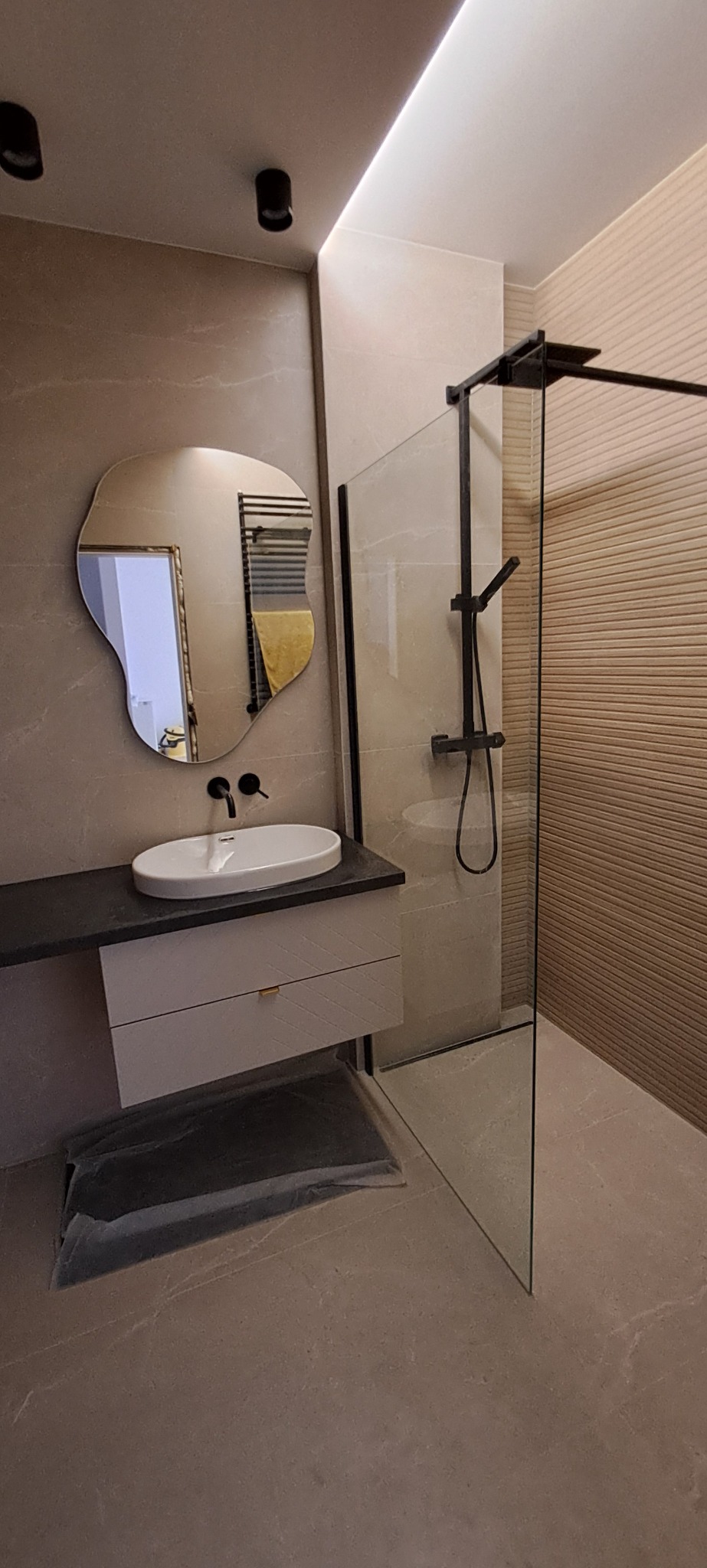 Nowoczesna łazienka z umywalką na szafce, lustrem o nieregularnym kształcie i kabiną prysznicową z czarnymi profilami. Beżowe płytki na ścianach i podłodze.