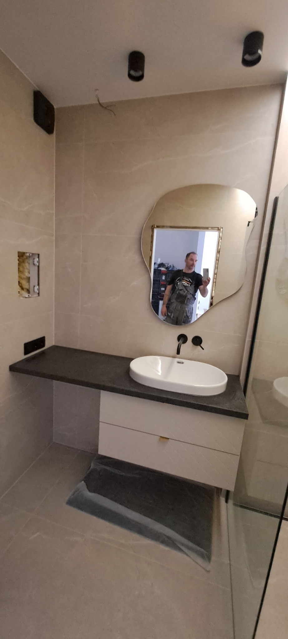 Wykończona łazienka z beżowymi płytkami, asymetrycznym lustrem i blatem z umywalką. Widoczne czarne baterie i oświetlenie sufitowe. W lustrze odbija się instalator.