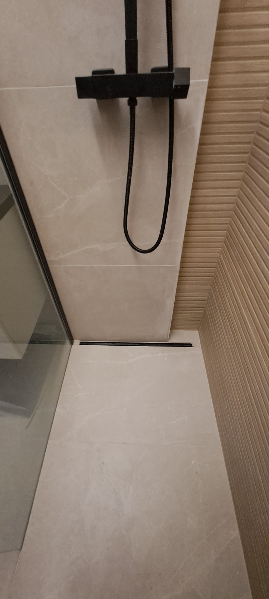 Nowoczesna kabina prysznicowa z czarną armaturą, beżowe płytki imitujące marmur i odpływ liniowy. Minimalistyczny design, eleganckie wykończenie.